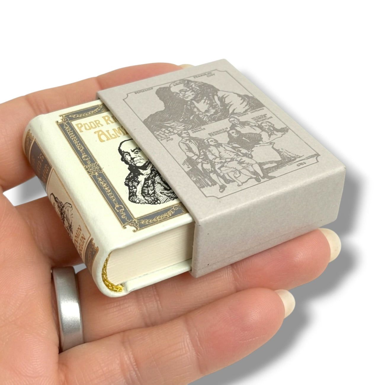 A miniature book-shaped matchbox or small box being held in a hand. The box is cream-colored with illustrated vintage book-style borders, a gray lid featuring black line drawings, and a small gold chain detail on the side.