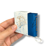 A hand holding a small packaged product, likely a matchbox-style box, with a white front panel featuring a gold eagle illustration and blue side panels. The product is photographed against a clean white background.