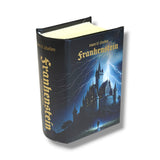 A hardcover book of Mary Shelley's 'Frankenstein' shown at a three-quarter angle on a white background. The cover features a dark blue illustration of a lightning-lit castle with a lone figure in the foreground, and the title is printed in gold gothic lettering.