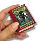 A hand holding a miniature book replica of 'The Legend of Silky Hollow' by Washington Irving. The book features a dark red cover with gold lettering and a black-and-gray illustration of a woman riding a wolf against a full moon.
