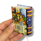 A hand holding a miniature hardcover book with an illustrated cover and purple spine, featuring the title 'Alice' and a barcode on the back.