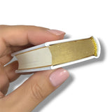 Hand holding a small book-shaped object with gold and white design on a white background