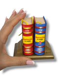 Two miniature books with colorful covers held by a hand on a white background. Mafalda