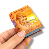 A hand holding a miniature book replica of 'O Pensamento Vivo de Mahatma Gandhi' against a clean white background.