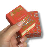 Hand holding a small red book with gold and blue floral designs on a white background. La Edad de la Inocencia