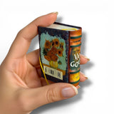 Hand holding a miniature book with Van Gogh's 'Sunflowers' design on a white background