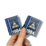 Two miniature books titled '1984' by George Orwell held by a hand on a white background.