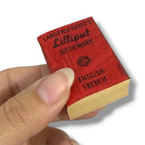 Miniature red book titled 'Langenscheidt's Lilliput Dictionary' held by a hand on a white background