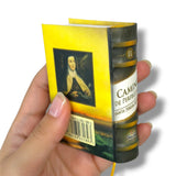 Hand holding a miniature book with a yellow cover and visible text and images.