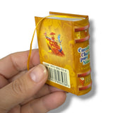 Hand holding a small, colorful book-shaped package with a visible barcode on a white background