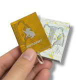 A hand holding a small yellow Hufflepuff-themed notebook in front of a matching patterned box on a white background.