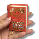Miniature red book with gold embossed design held in a hand against a white background. La Edad de la Inocencia