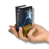 Hand holding a miniature book titled 'Frankenstein' with a wooden stand on a white background