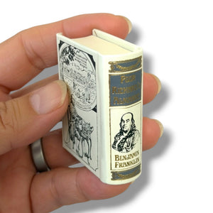 A miniature replica of 'The Diary of Benjamin Franklin' held in a person's hand against a white background.