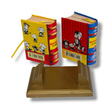 Two miniature books with colorful covers on a gold stand against a white background Mafalda