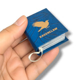 A miniature blue book with a gold Ravenclaw eagle logo and the word 'RAVENCLAW' on the cover, held in a person's hand against a white background.