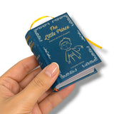 A hand holding a miniature book edition of 'The Little Prince' with a dark blue cover, gold lettering, and a yellow ribbon bookmark, set against a plain white background.
