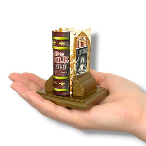 Miniature book and cover displayed on a stand held in a hand against a white background. La Sacrée Bible Illustrée