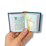 A hand holds a miniature open book with colorful striped pages, a blue cover edge, and a yellow ribbon bookmark against a plain white background. The Little Prince