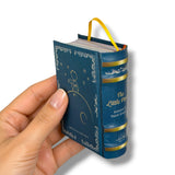 A hand holding a miniature blue book styled like The Little Prince, with gold lettering and trim, a yellow ribbon bookmark, and white page edges visible against a white background.