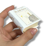 Hand holding a miniature book titled 'The Little Prince' with a cat illustration on the cover.