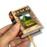A miniature book-shaped bookmark or pendant held in a hand. The cover features a scenic image of Machu Picchu with the title 'CUSCO Ombigo del mundo' and the brand 'Aventura Criativa'.