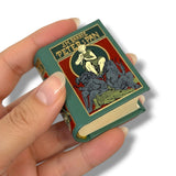 Hand holding a miniature book with a colorful cover featuring text and illustrations.