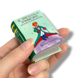 A hand holding a miniature book of Il Piccolo Principe (The Little Prince) with a light green cover and green spine, set against a plain white background.