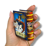 Hand holding a small, colorful book-shaped object with a cartoon character and 'Magic' text on a white background.