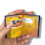 Hand holding a small open book with colorful illustrations and text. Magia para Niños