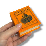 Hand holding a miniature book titled 'The Complete Plays' by William Shakespeare with an orange cover.