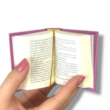 Hand holding a small open book with purple edges on a white background.Sentido y Sensibilidad