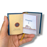 A hand holding a miniature open book with blue covers and cream-colored pages. The left page shows a decorative circular emblem with the text 'Illustrations by the Author,' and the right page shows the heading 'Chapter 1' above a small illustration. The little Prince