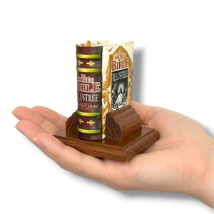 Miniature book and candle set on a wooden stand held in a hand against a white background. La Sacrée Bible Illustrée