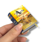 Hand holding a miniature book titled 'El Arte de la Guerra' with a yellow and blue cover.