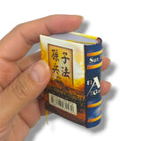 Hand holding a miniature book titled 'Sun Tzu' on a white background