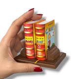 Hand holding a miniature book set on a wooden stand with colorful covers. Condorito