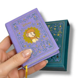Hand holding a small purple book with a character illustration on a white background. Bernardo y Bianca