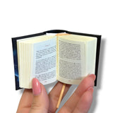 A miniature open book bookmark held in a hand. The tiny book has a black cover, cream-colored pages, and a small gold ribbon bookmark, shown against a plain white background. Frankenstein 
