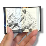 A hand holding a miniature sketchbook with a detailed black and white sketch of mountains on the pages.