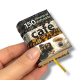 A hand holding a miniature book titled '150 maneras de disfrutarlo café' with a coffee-themed cover, featuring coffee beans, a cup of coffee, and a yellow bookmark ribbon, set against a plain white background.