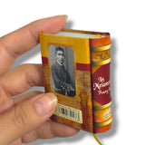 Miniature book with a person's portrait on the cover held by a hand against a white background. La Metamorfosis