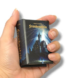 A hand holding a miniature book-shaped product featuring Mary Shelley's Frankenstein, with a dark cover illustration of a castle under lightning and gold title text.