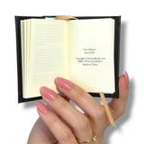 A hand with pink manicured nails holds a miniature black book Frankenstein 
 with cream-colored pages open, showing publication details on the right page.