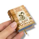 A miniature book-shaped product held in a person's hand against a clean white background. The item is designed to look like a small vintage book with a wood-grain cover, Portuguese text reading 'O Professor O Loco A Procissão' and 'Kahlil Gibran', a sepia portrait illustration, and a blue ribbon bookmark.