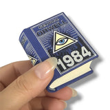 Miniature book of '1984' by George Orwell held by a hand on a white background