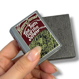 A hand holds a small square book or card titled "Henry James The Turn of the Screw" with a gray cover, next to the textured gray back of another matching item on a white background.