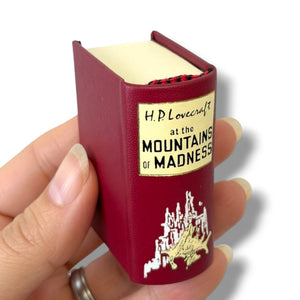A hand holding a miniature book-shaped item featuring the cover art and title H.P. Lovercraft. At the Montains of Madness'H.P.