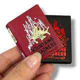 A hand holds two miniature book-shaped items featuring H.P. Lovercraft. At the Montains of Madness