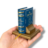 A miniature book-shaped bookend holding 'The Little Prince' on a gold-toned base, shown resting on a person's hand against a white background.
